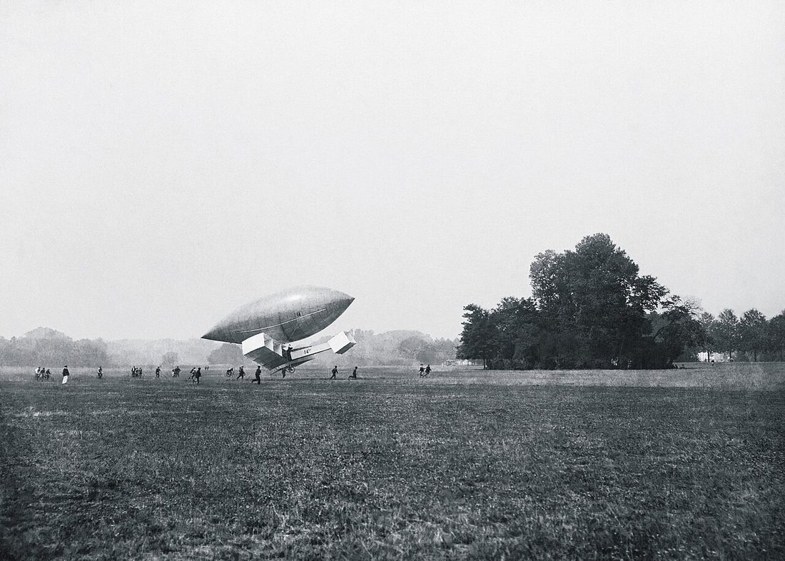 Campo aberto com o avião 14-bis suspenso sob um dirigível alongado, cercado por pessoas, em fotografia antiga em preto e branco.