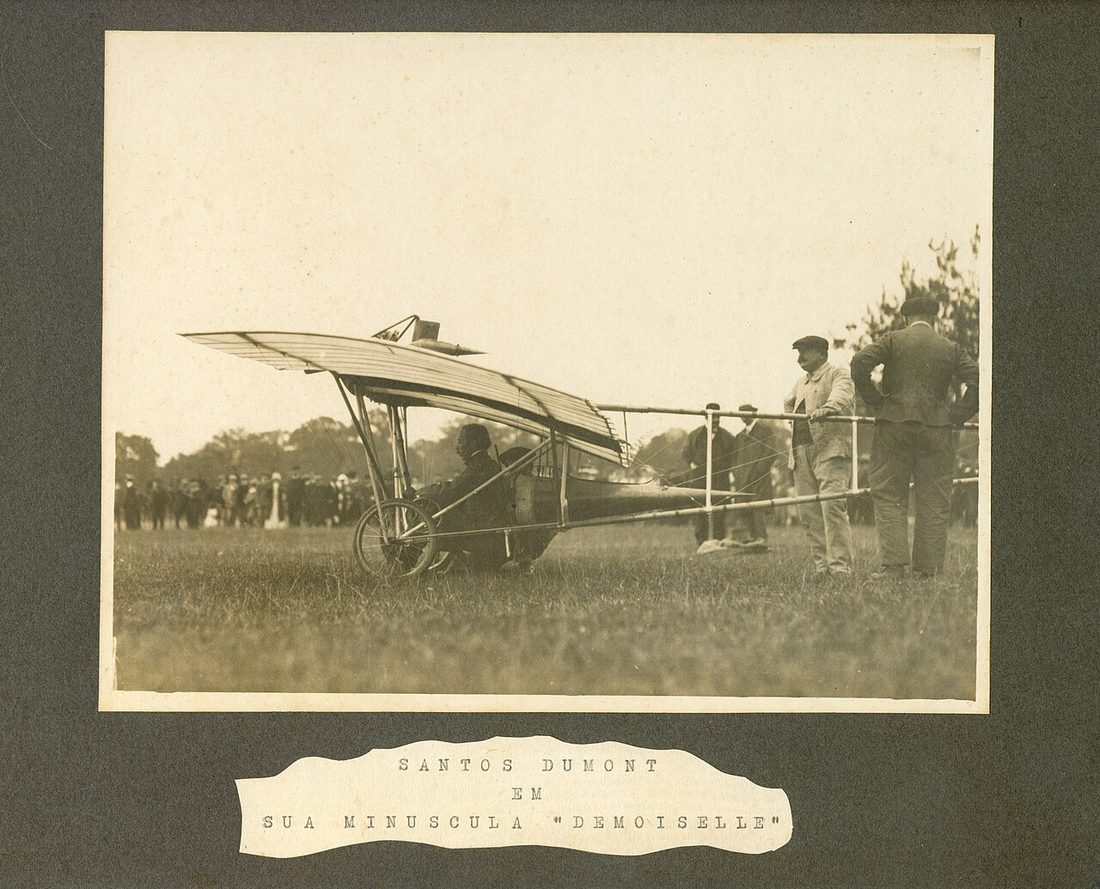 Fotografia antiga montada em cartão, com o pequeno monoplano Demoiselle no campo e homens em volta.