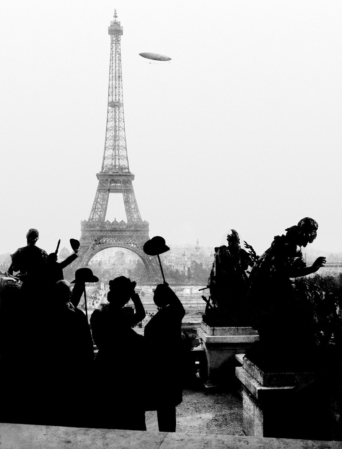 Vista do Trocadéro com a Torre Eiffel ao fundo e o dirigível número 6 no céu, cercado por silhuetas de pessoas observando.