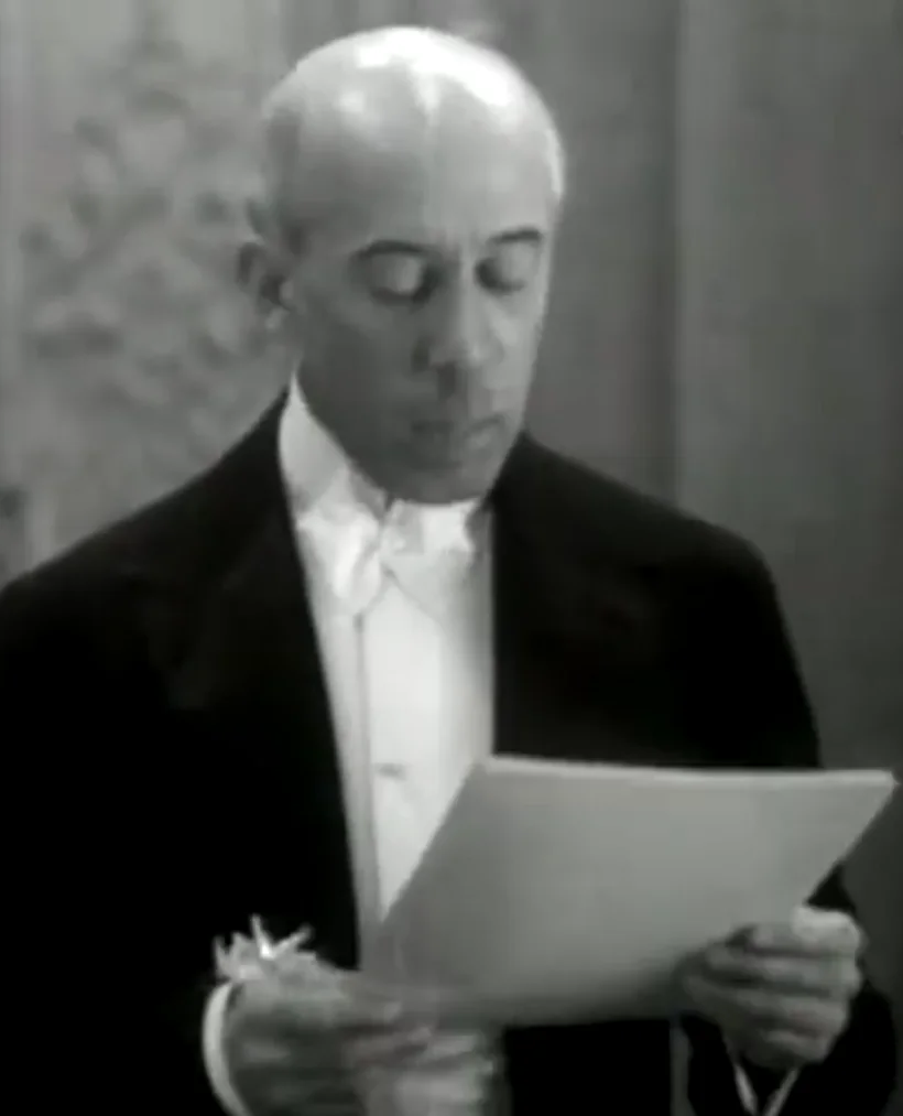 Santos Dumont, de casaca, lendo uma folha em voz alta